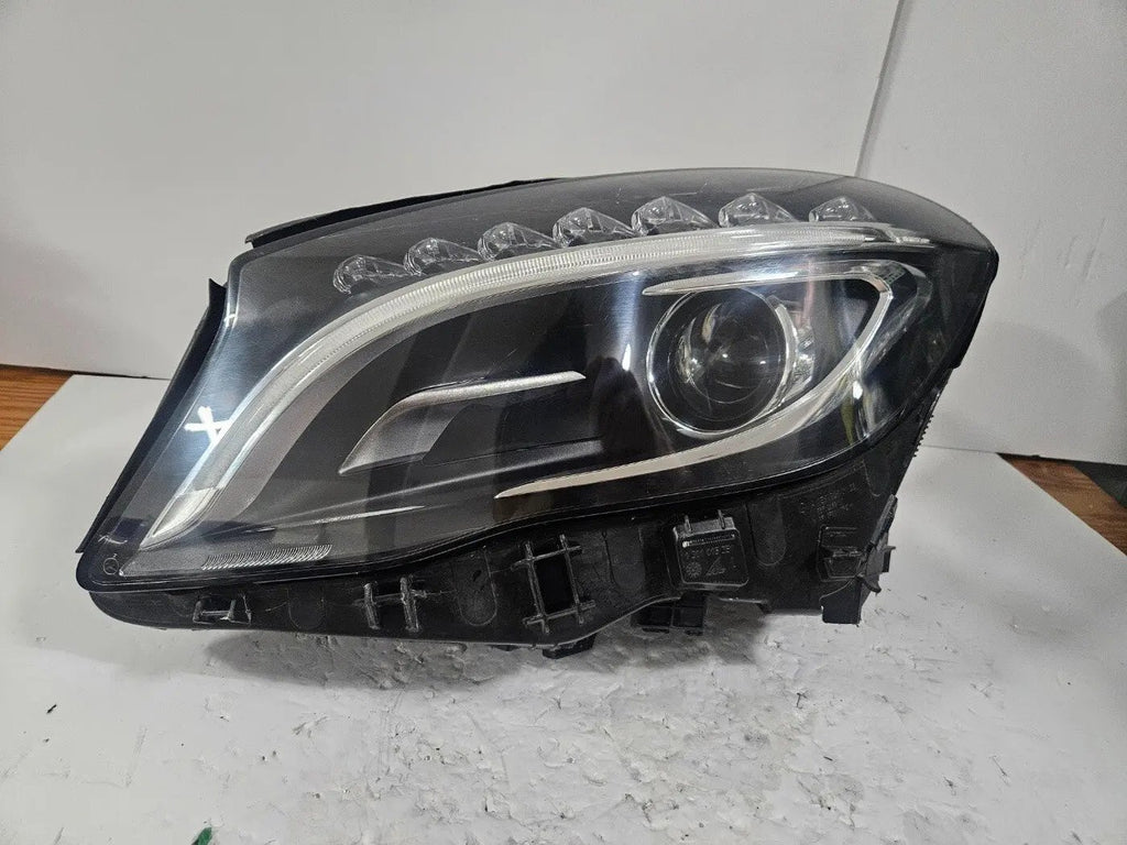Genuine used Mercedes-Benz Bi-Xenon headlight for GLA-Class model X156. This left-side (LH) OEM headlamp, part number A1569061700, features Bi-Xenon lighting technology for enhanced night visibility and a focused beam pattern. The image shows the original left headlight in used condition, highlighting the lens clarity, projector unit, mounting brackets, and electrical connector — ideal for replacing a damaged headlamp on compatible Mercedes-Benz GLA X156 vehicles.

